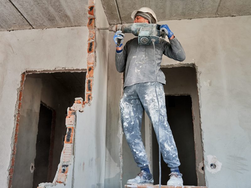 Full length of man in workwear drilling wall with hammer drill. Male worker using drill breaker while destroying wall in apartment under renovation. Demolition work and home renovation concept.